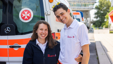 Zwei Ehrenamtliche Arm in Arm vor einem Rettungswagen.