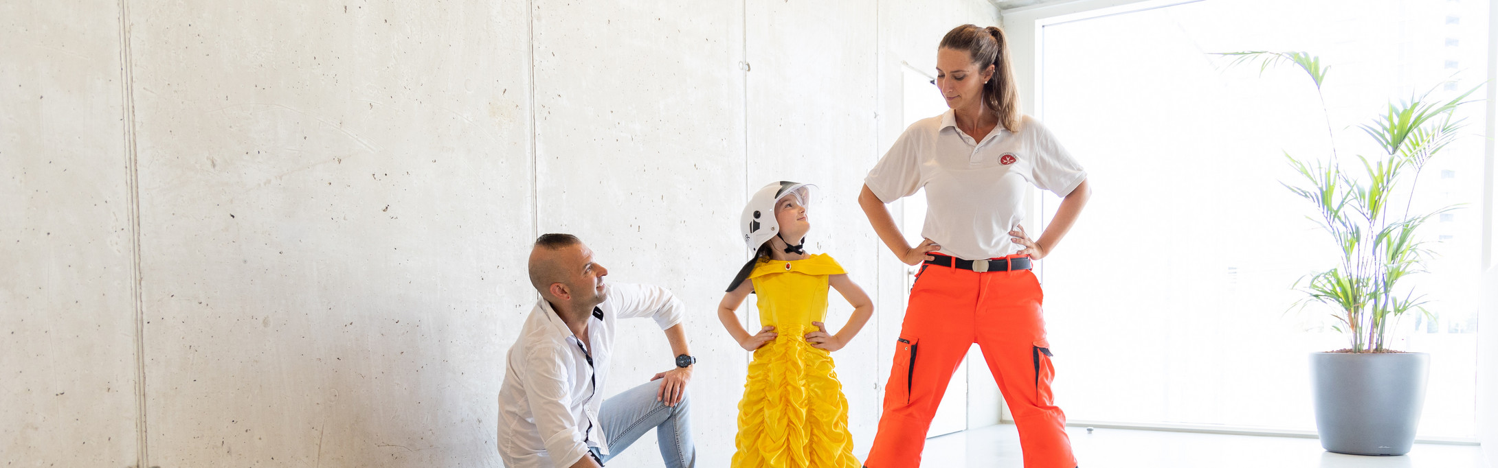 Ein Mann, der am Boden kniet, und ein Mädchen, das ein gelbes Prinzessinenkleid und einen Feuerwehrhelm trägt, schauen zu einer Johanniterin auf, die in einer Superhelden-Pose neben ihnen steht.